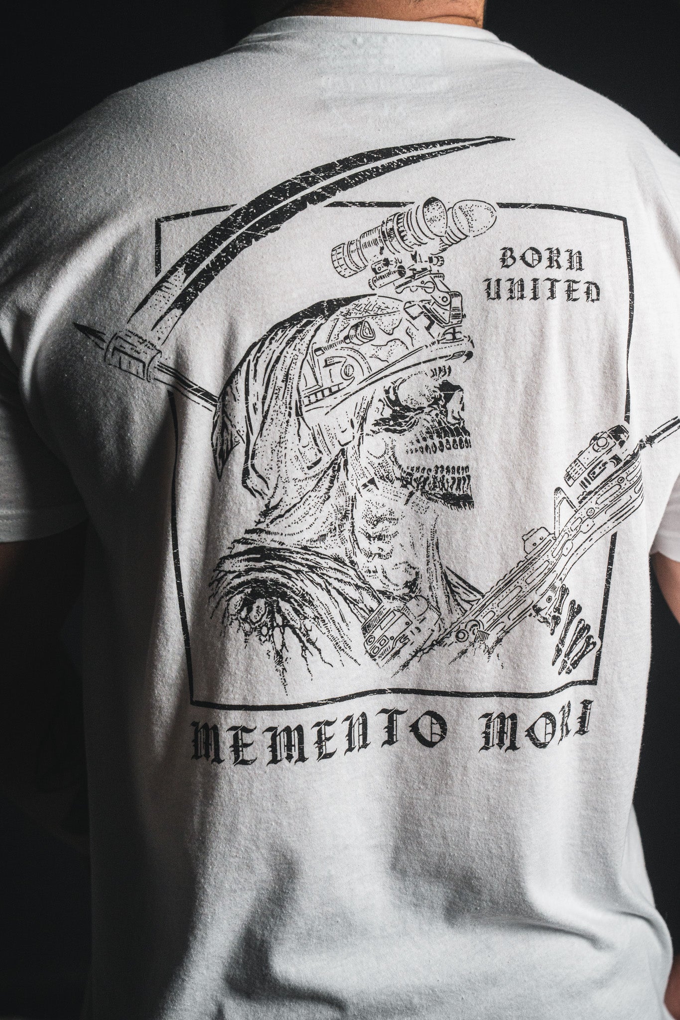 A person wearing the Memento Mori - White t-shirt from Born United Private Label, featuring a detailed skeletal graphic with “Born United” above and “Memento Mori” below—a philosophical reminder of our shared mortality.