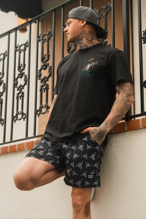 A tattooed man in a black t-shirt and Relentless Betrayal Mens Performance Athletic Shorts | 5.5- Sink or Swim, wearing a backward cap, leans against a white wall by a decorative metal railing with one hand in his pocket.