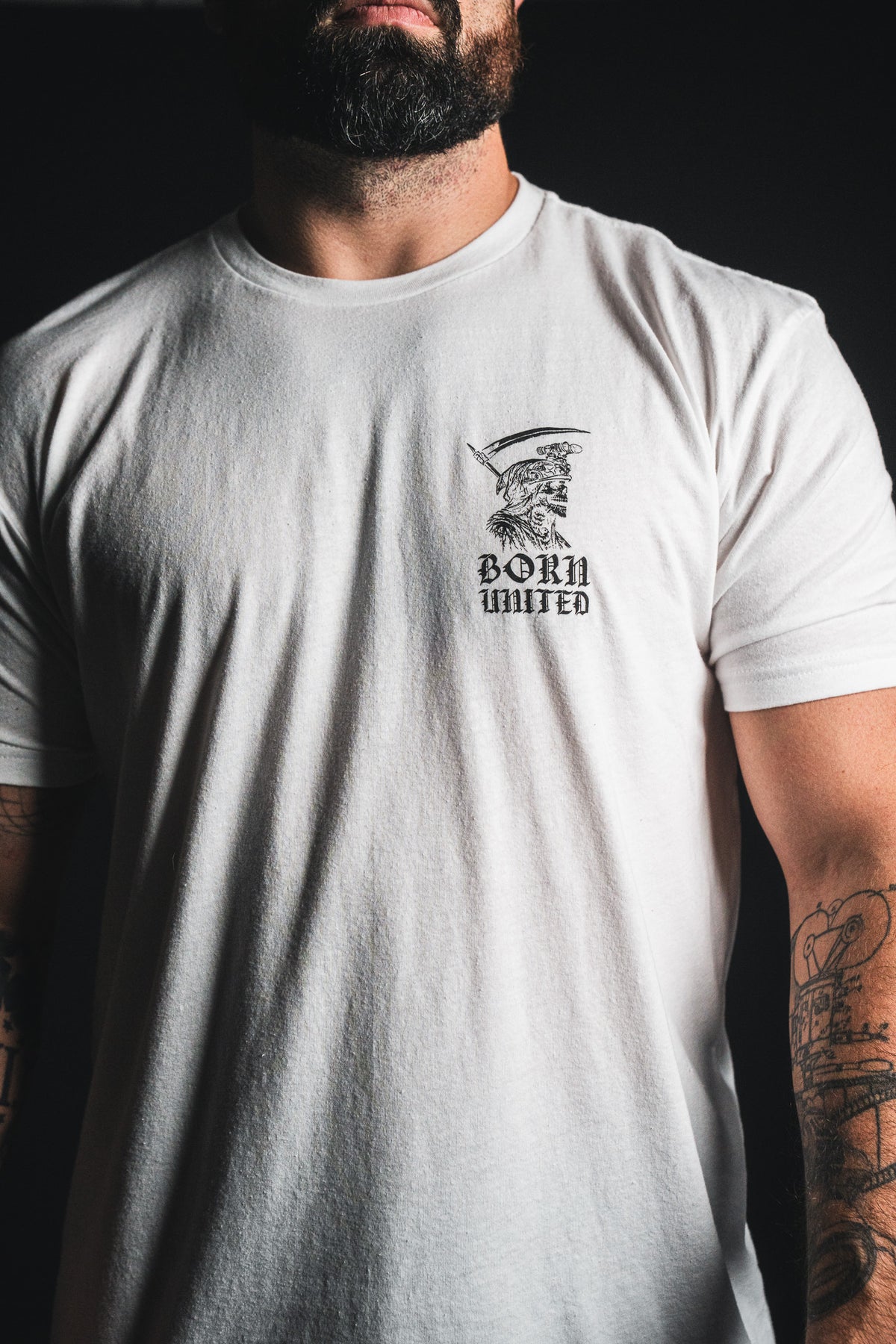 A bearded man wears the “Memento Mori - White” t-shirt from Born United Private Label, which features a small left-chest graphic of a skull and helmet—a subtle memento mori, thoughtfully reminding us of our shared mortality. His tattooed arms are partially visible.