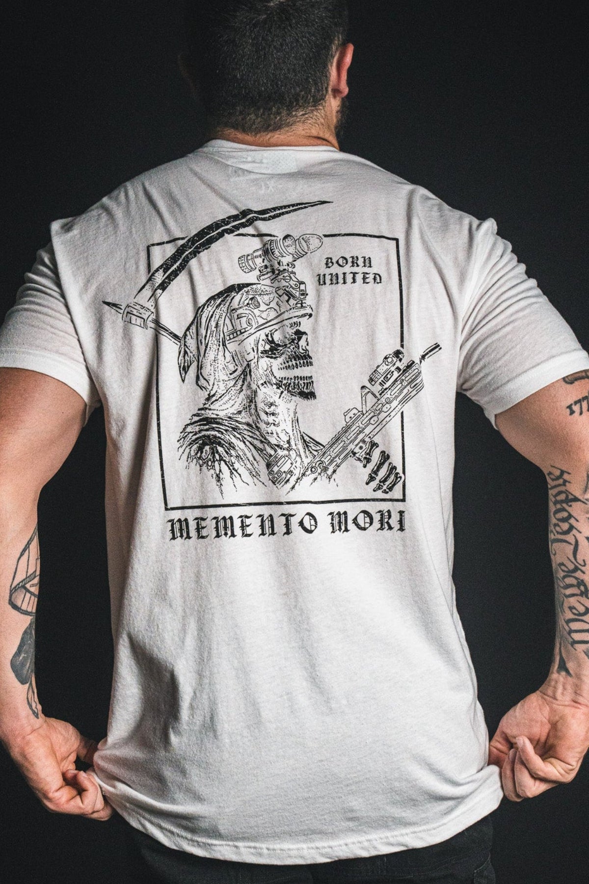 A person wearing the Born United Private Label “Memento Mori - White” T-shirt, featuring a skull graphic, crossed weapons, and the text Born Hunted and Memento Mori—a philosophical reminder of mortality—on the back, standing against a dark background.