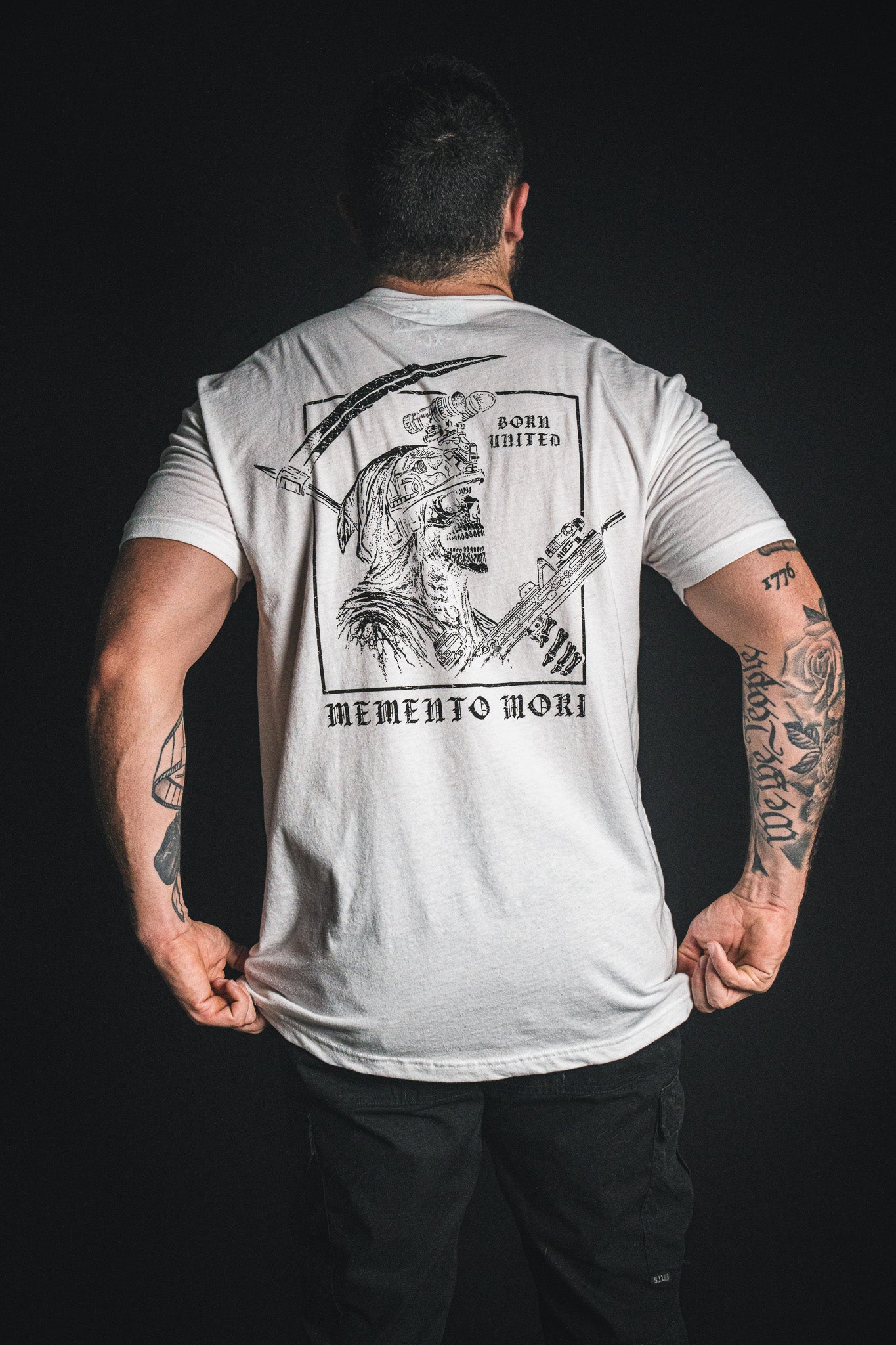 A person wearing the Born United Private Label “Memento Mori - White” T-shirt, featuring a skull graphic, crossed weapons, and the text Born Hunted and Memento Mori—a philosophical reminder of mortality—on the back, standing against a dark background.