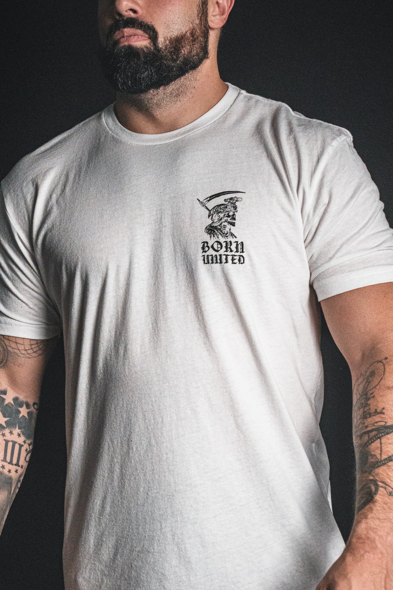 A man with tattoos wears the Born United Private Label “Memento Mori - White” t-shirt, featuring a small illustrated graphic on the upper left chest—a subtle philosophical reminder of mortality seamlessly integrated into his everyday style.