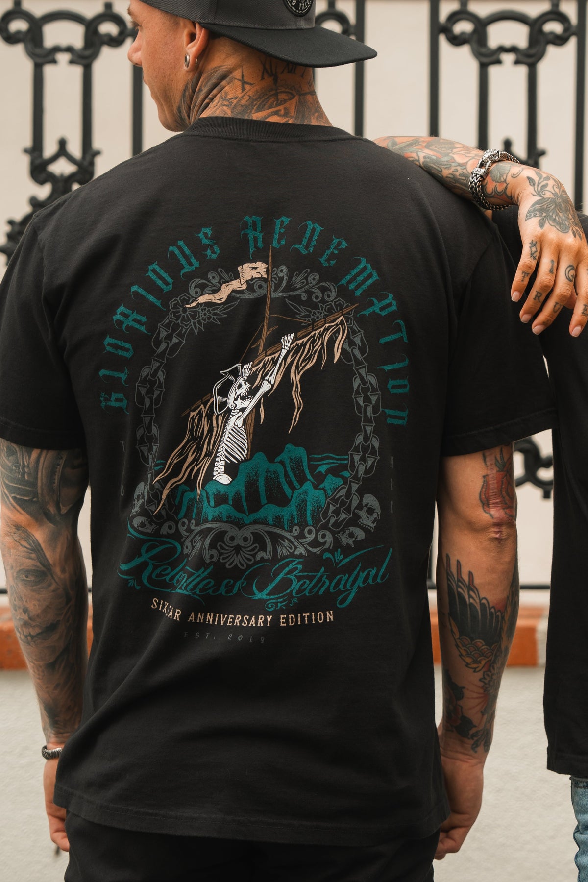 A tattooed individual is wearing the Relentless Betrayal Glorious Redemption Premium Tee, a black jersey cotton T-shirt featuring a graphic design and the text “Road to Redemption” and “Reader of Betrayal 6th Anniversary Limited Edition.”.