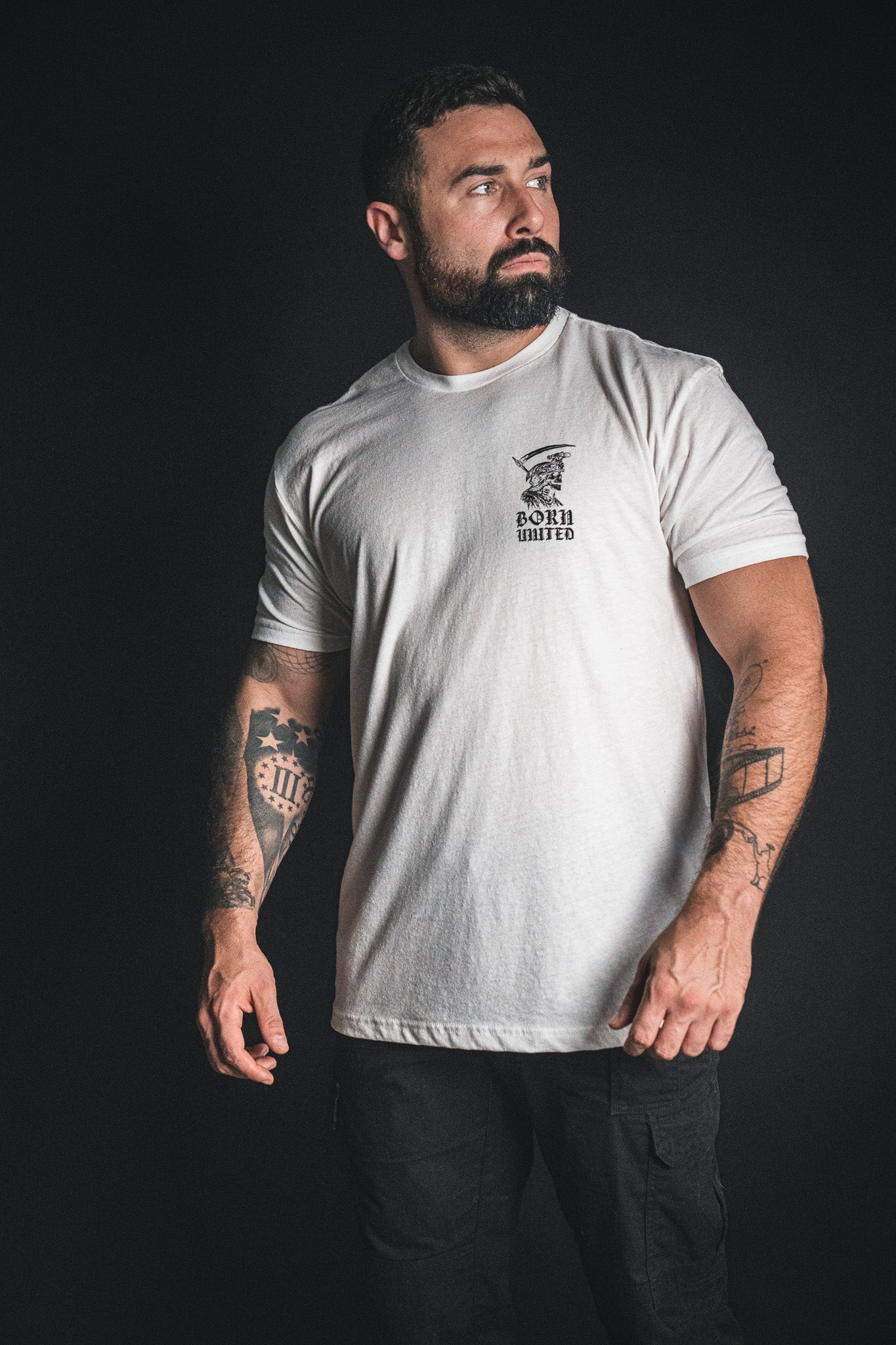 A bearded man with tattoos wears the Born United Private Label “Memento Mori - White” t-shirt and black pants, standing against a dark background and gazing to his left—a subtle but powerful reminder of mortality inspired by the philosophy behind memento mori.