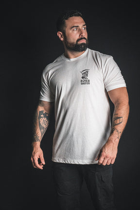 A bearded man with tattoos wears the Born United Private Label “Memento Mori - White” t-shirt and black pants, standing against a dark background and gazing to his left—a subtle but powerful reminder of mortality inspired by the philosophy behind memento mori.