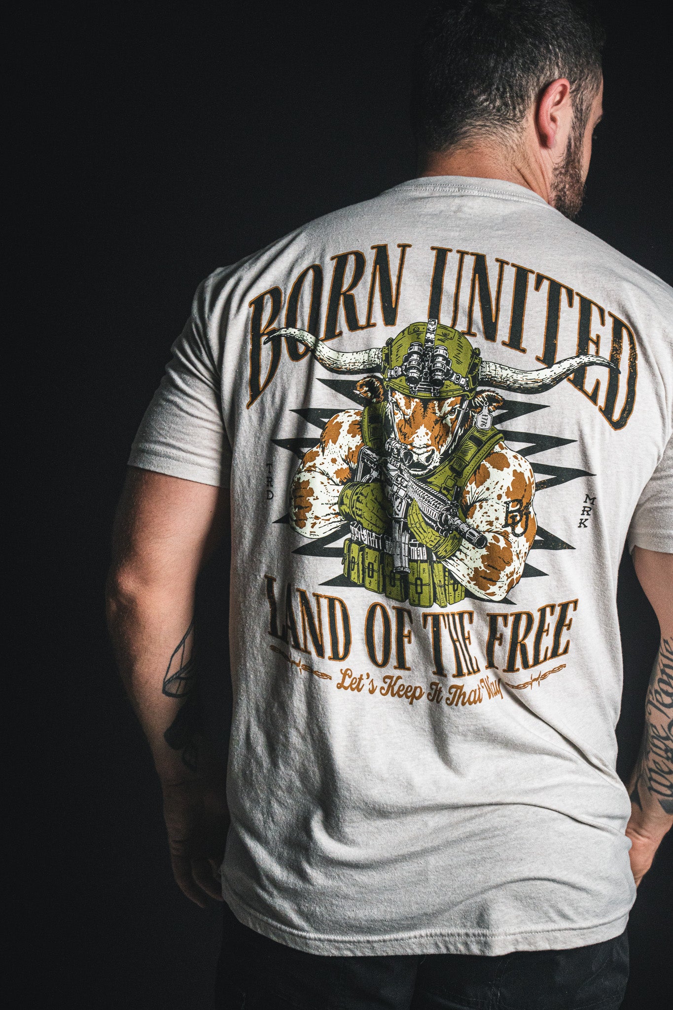 A man is shown from the back, wearing the Born United Private Label “Freedom Frontier” T-shirt—a light gray tee featuring a bold bull skull graphic with American flag motifs and the words “Born United, Land of the Free” on the back, celebrating American values and patriotism.
