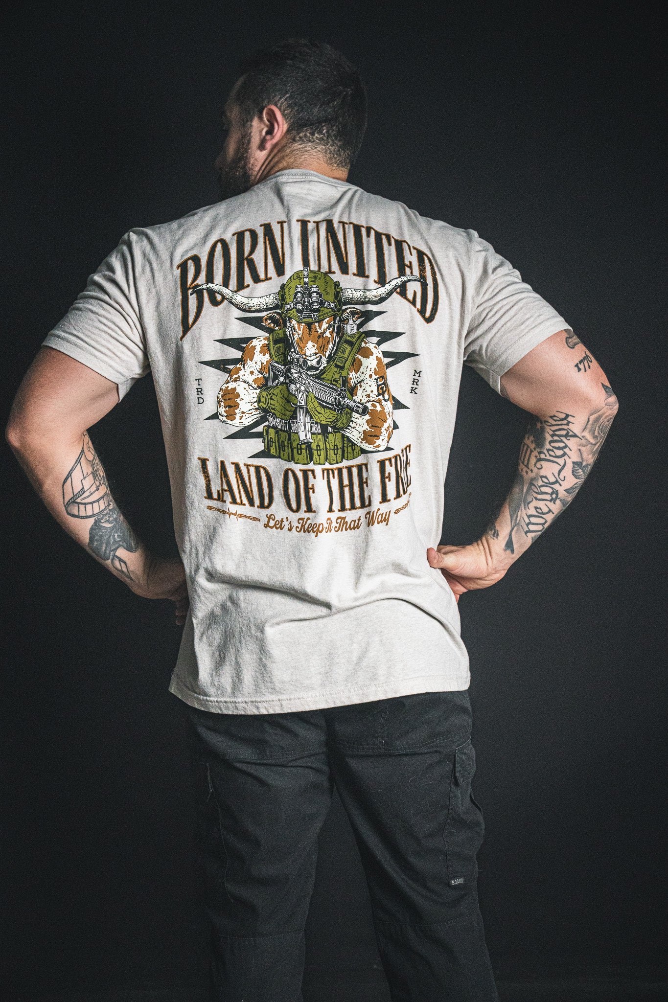 A man with tattooed arms stands against a dark background, wearing the Freedom Frontier light t-shirt from Born United Private Label, featuring an American flag eagle design and “BORN UNITED LAND OF THE FREE”—a bold statement of freedom and American pride.