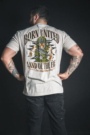 A man with tattooed arms stands against a dark background, wearing the Freedom Frontier light t-shirt from Born United Private Label, featuring an American flag eagle design and “BORN UNITED LAND OF THE FREE”—a bold statement of freedom and American pride.