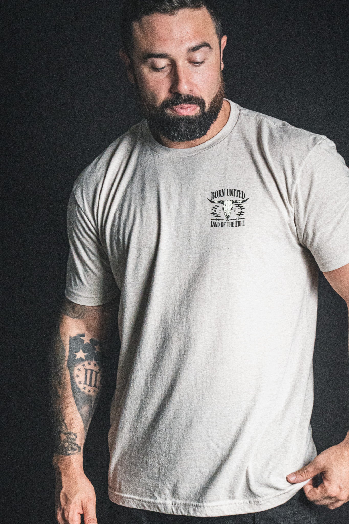 A bearded, tattooed man stands against a dark background, eyes closed and head bowed, wearing a light-colored Born United Private Label Freedom Frontier t-shirt—a quiet moment embodying American values and freedom.