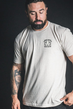 A bearded, tattooed man stands against a dark background, eyes closed and head bowed, wearing a light-colored Born United Private Label Freedom Frontier t-shirt—a quiet moment embodying American values and freedom.
