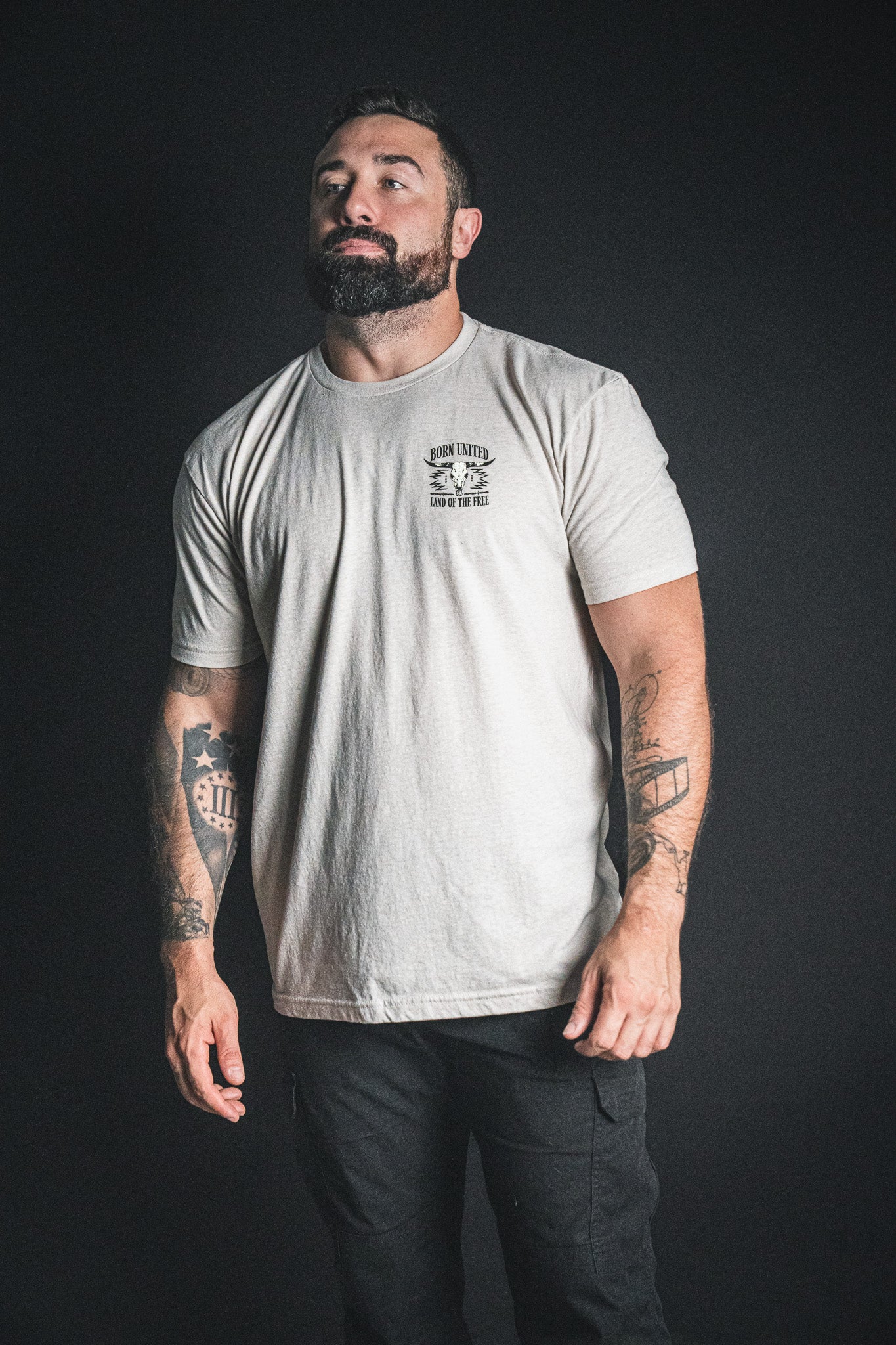 A bearded man with tattoos, dressed in Born United Private Label’s Freedom Frontier light-colored t-shirt and black pants, stands against a dark background, gazing upward and to the side—capturing the quiet spirit of freedom.