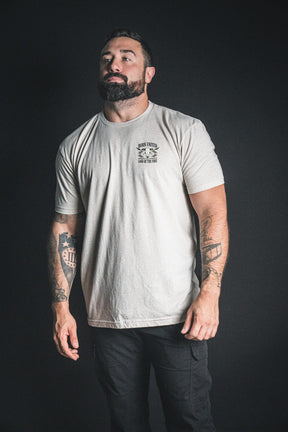 A bearded man with tattoos, dressed in Born United Private Label’s Freedom Frontier light-colored t-shirt and black pants, stands against a dark background, gazing upward and to the side—capturing the quiet spirit of freedom.