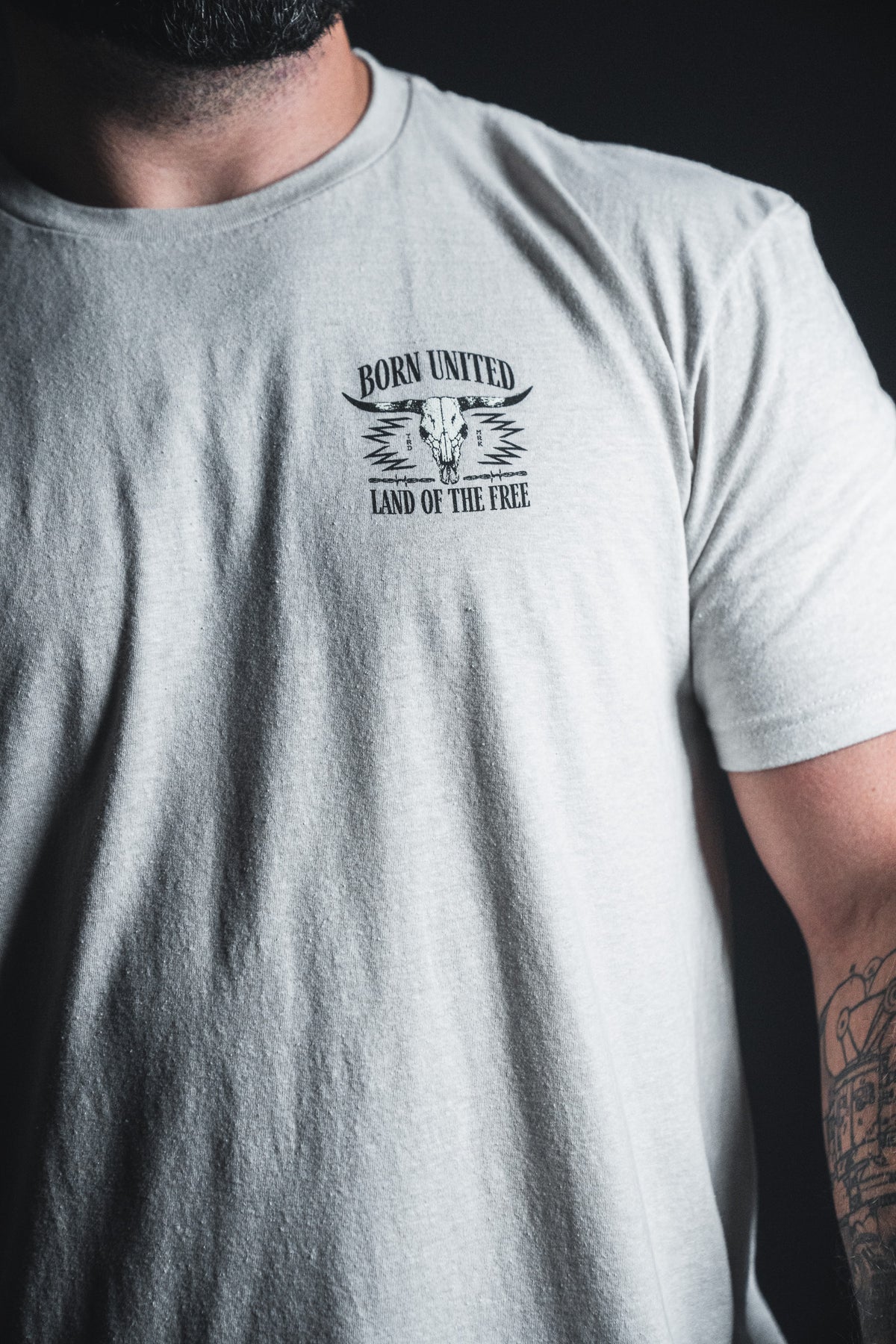 A person is wearing the Freedom Frontier T-shirt from Born United Private Label, featuring a skull graphic and “Land of the Free” design in light gray—a patriotic piece that reflects American values; part of their tattooed arm is visible.