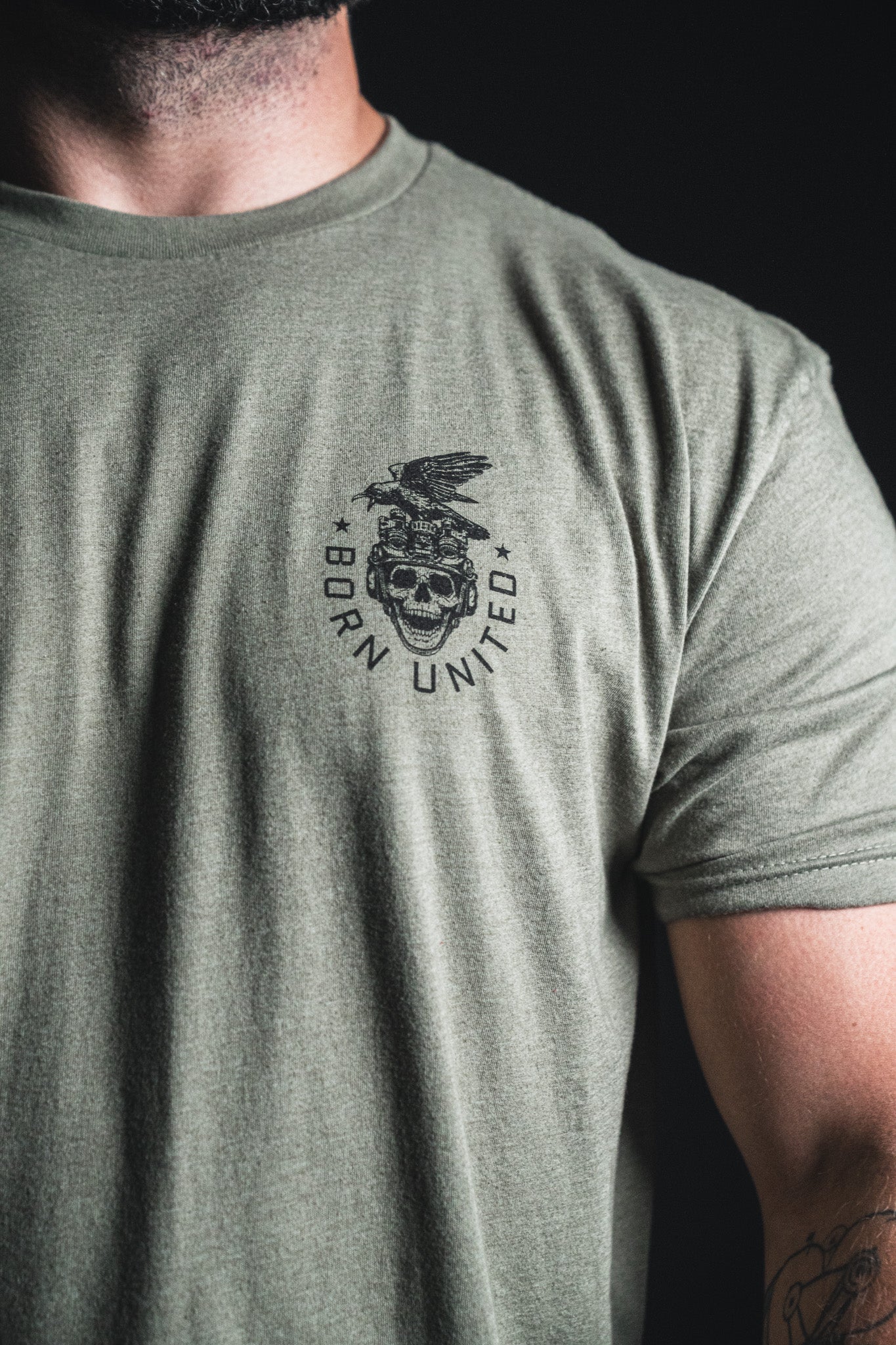 A person wearing an olive green “Only the Dead” t-shirt from Born United Private Label, featuring a skull, eagle, stars, and the words Born United on the left chest—echoing themes of remembrance and history.