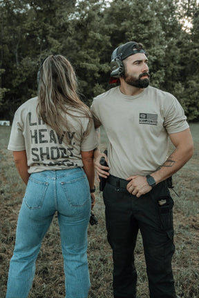 A woman and a man stand on grass, both wearing the LHSG - Tan Tee from Tango Charlie Apparel. They look relaxed with trees in the background.