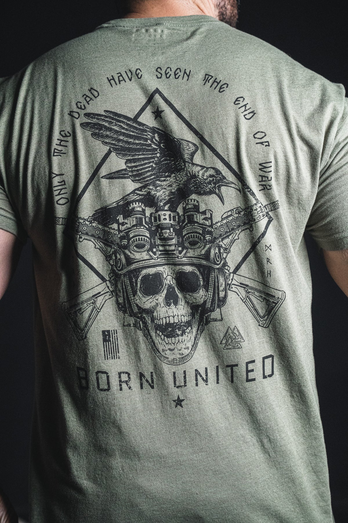 Wearing the “Only the Dead” green T-shirt from Born United Private Label, a person sports a graphic featuring a skull, crossed rifles, night vision goggles, an eagle, and the war quote “Only the dead have seen the end of war”—famously attributed to George Santayana—along with the Born United brand phrase.
