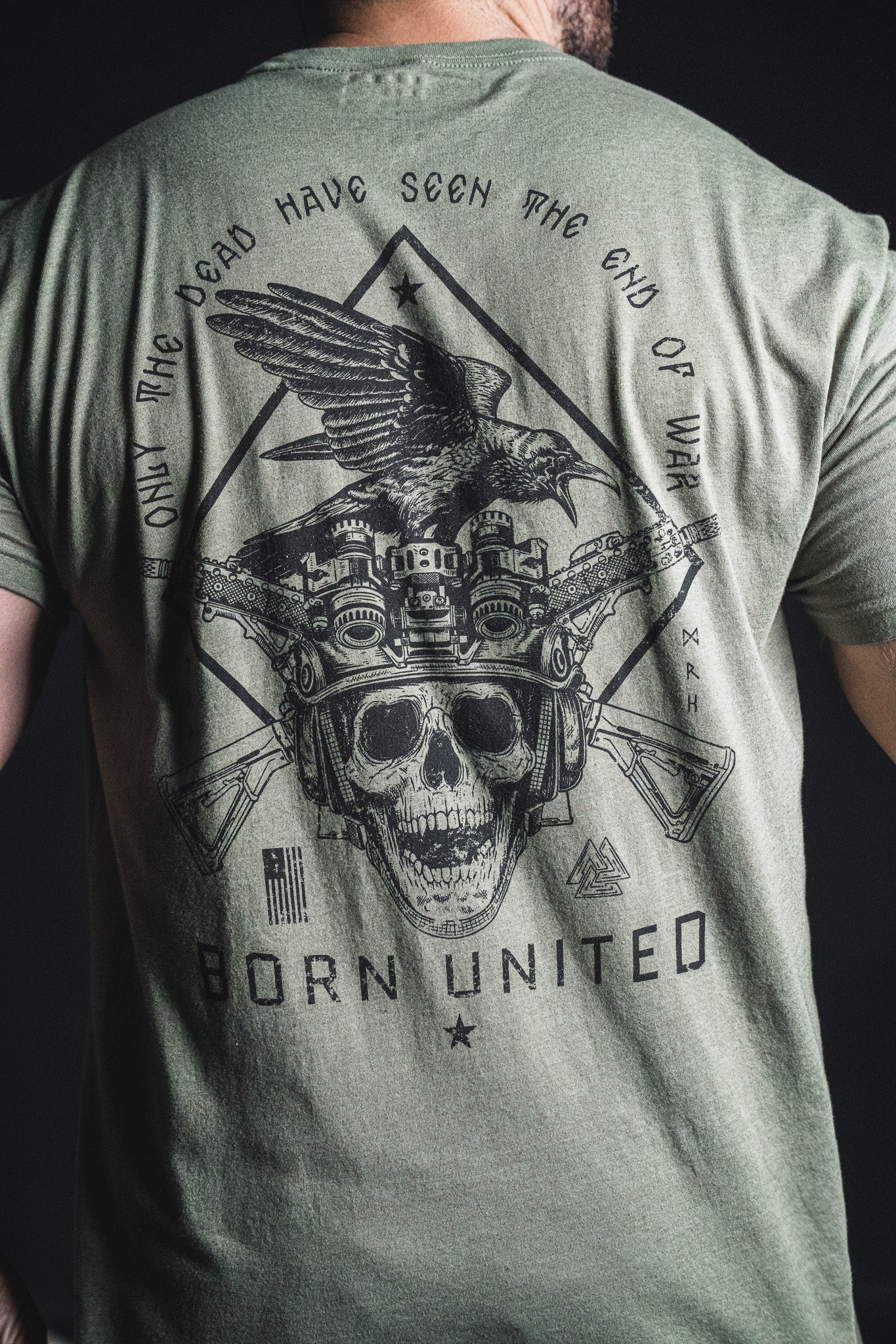 Wearing the “Only the Dead” green T-shirt from Born United Private Label, a person sports a graphic featuring a skull, crossed rifles, night vision goggles, an eagle, and the war quote “Only the dead have seen the end of war”—famously attributed to George Santayana—along with the Born United brand phrase.