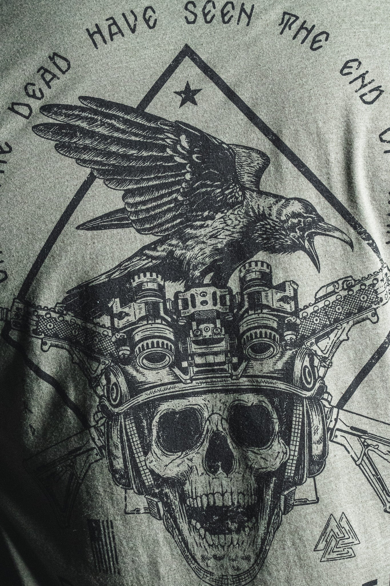A black graphic on gray fabric features a crow perched atop a skull wearing night vision goggles, encircled by partial text inspired by a George Santayana war quote; this design is titled “Only the Dead” from the Born United Private Label.