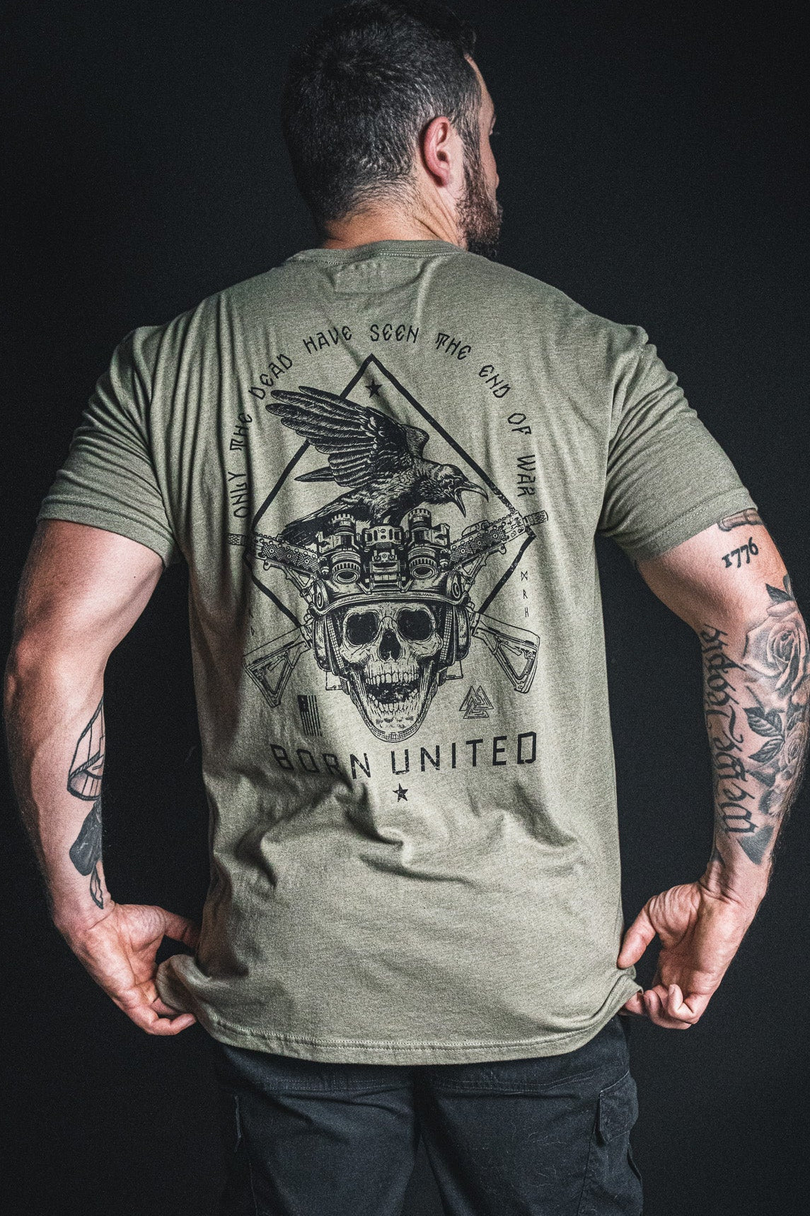 A man stands with his back to the camera, wearing an olive green “Only the Dead” t-shirt by Born United Private Label, featuring a bold graphic with a skull, eagle, crossed rifles, and the brand’s name. His tattooed arms are visible below the sleeves. The design evokes classic wartime quotes and echoes George Santayana’s famous reflections on conflict.
