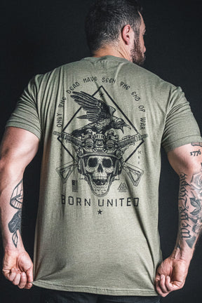 A man wears the “Only the Dead” olive green t-shirt by Born United Private Label, featuring a striking back design with a skull, crow, rifles, tanks, and the powerful George Santayana war quote: “Only the dead have seen the end of war.”.