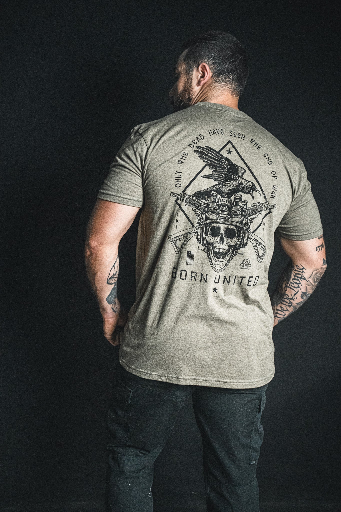 A man stands with his back to the camera, wearing the “Only the Dead” T-shirt from Born United Private Label, which features a striking design of a skull, eagle, and guns alongside George Santayana’s iconic quote—“Only the dead have seen the end of war”—and bold “Born United” text.