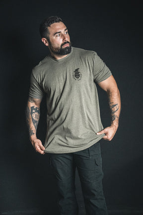 A man with tattoos wears the “Only the Dead” khaki t-shirt by Born United Private Label and black pants, standing against a dark background as he looks slightly to the side, holding the hem of his shirt; his quiet resilience calls to mind a famous George Santayana war quote.
