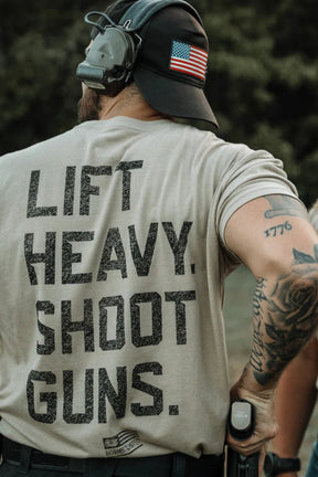 Wearing the Tango Charlie Apparel LHSG - Tan Tee, a person with tattoos on their right arm and a holstered handgun on their belt stands outdoors in high-intensity fitness gear and a backward cap with a US flag patch.