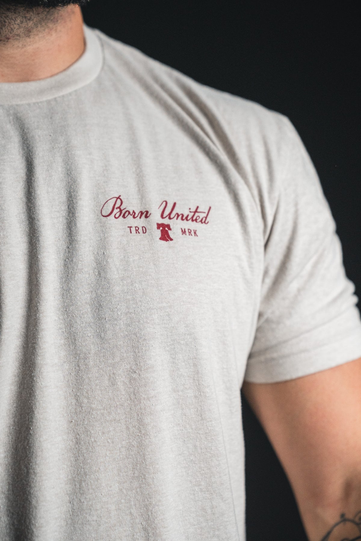 A person wears the Freedom Not Silenced light grey t-shirt by Born United Private Label, featuring “Born United TRD MRK” and a small red logo on the left chest, standing against a dark background—a subtle expression of freedom through minimalist style.