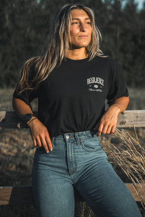 A woman with long hair stands by a wooden fence outdoors, wearing the Resilience - Premium Tee from Tango Charlie Apparel and high-waisted blue jeans, gazing confidently into the sunlight.
