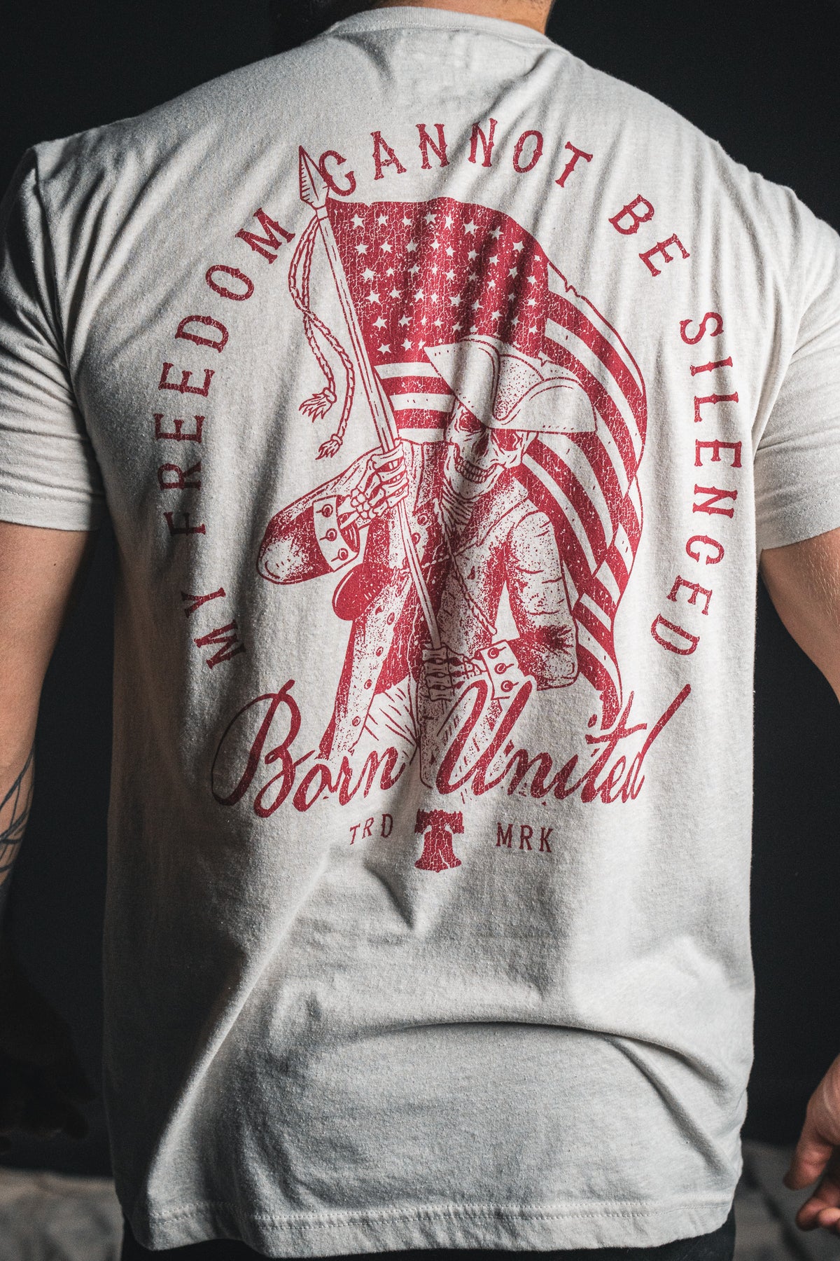 A person wearing the “Freedom Not Silenced” white T-shirt by Born United Private Label, featuring a graphic of a colonial soldier holding an American flag and the text “MY FREEDOM CANNOT BE SILENCED,” celebrating the enduring spirit of freedom, with “Born United” printed on the back.