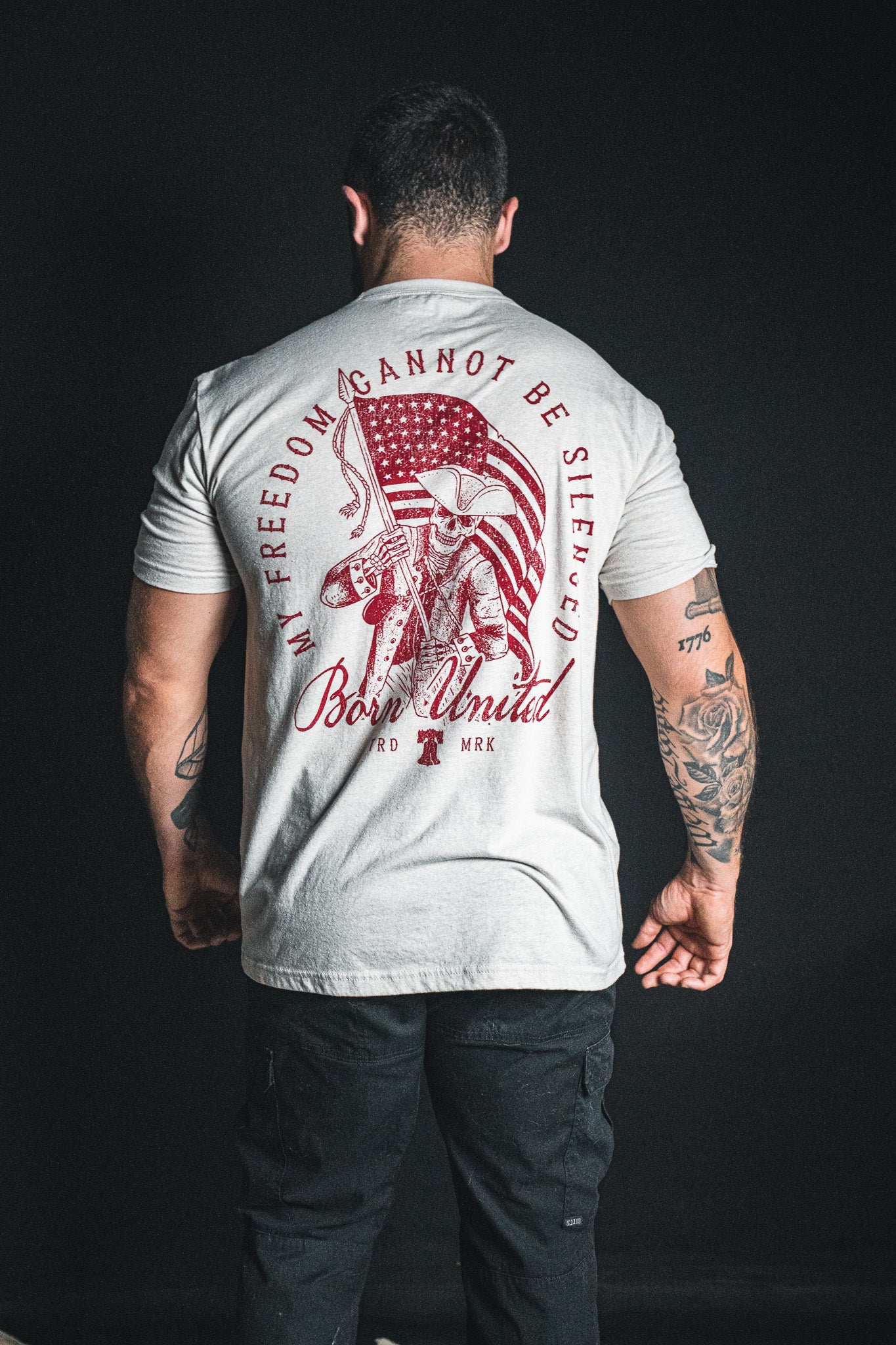 A man stands with his back to the camera, wearing the “Freedom Not Silenced” shirt by Born United Private Label. The bold graphic on his shirt shows a colonial figure raising a flag, accompanied by the powerful message: MY FREEDOM CANNOT BE SILENCED. His tattooed arms further showcase his unwavering dedication to freedom.