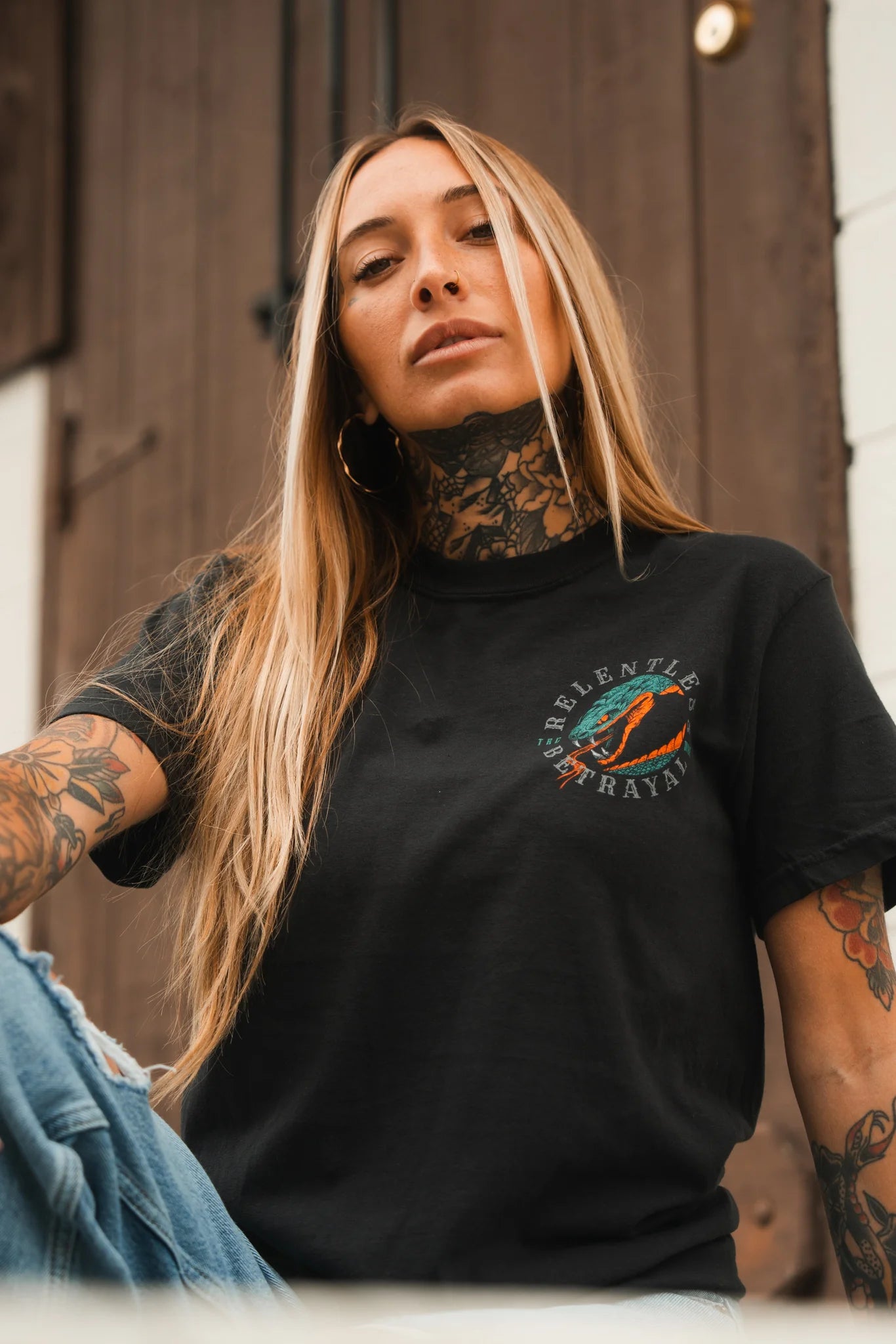 A woman with long blonde hair and visible tattoos wears a brand new black Don't Tread Here Premium Tee by Relentless Betrayal, featuring a colorful lips graphic, as she sits confidently in front of a wooden door, looking at the camera.