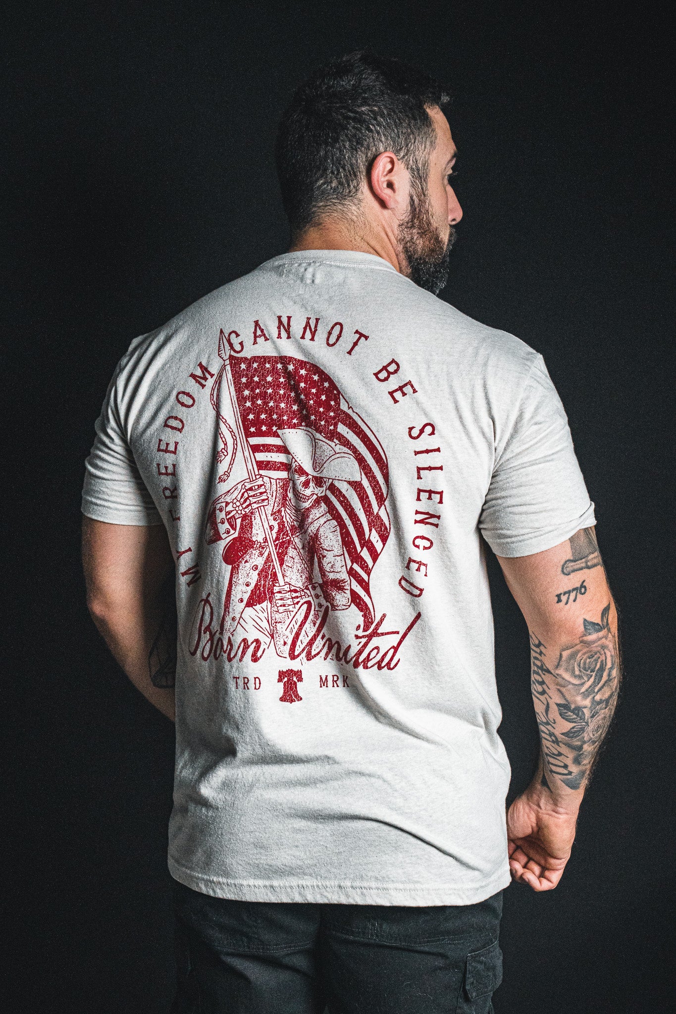 A man stands with his back to the camera, wearing a light-colored ‘Freedom Not Silenced’ T-shirt from Born United Private Label, featuring a graphic of a soldier holding an American flag and boldly conveying the unyielding spirit of freedom.