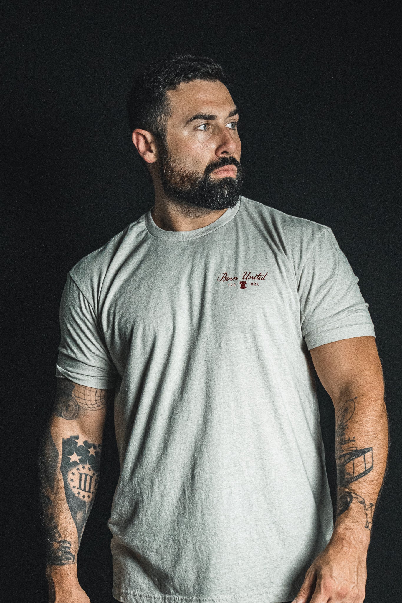 A bearded, tattooed man wears the Born United Private Label “Freedom Not Silenced” light gray t-shirt, standing against a dark background and gazing to the side as if contemplating freedom in a silenced world.