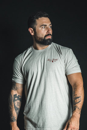 A bearded, tattooed man wears the Born United Private Label “Freedom Not Silenced” light gray t-shirt, standing against a dark background and gazing to the side as if contemplating freedom in a silenced world.