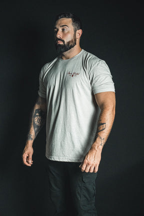 A man with a beard and tattoos stands against a dark background, wearing the Born United Private Label “Freedom Not Silenced” light gray T-shirt and dark pants, his sideways glance quietly hinting at freedom even in moments that feel silenced.