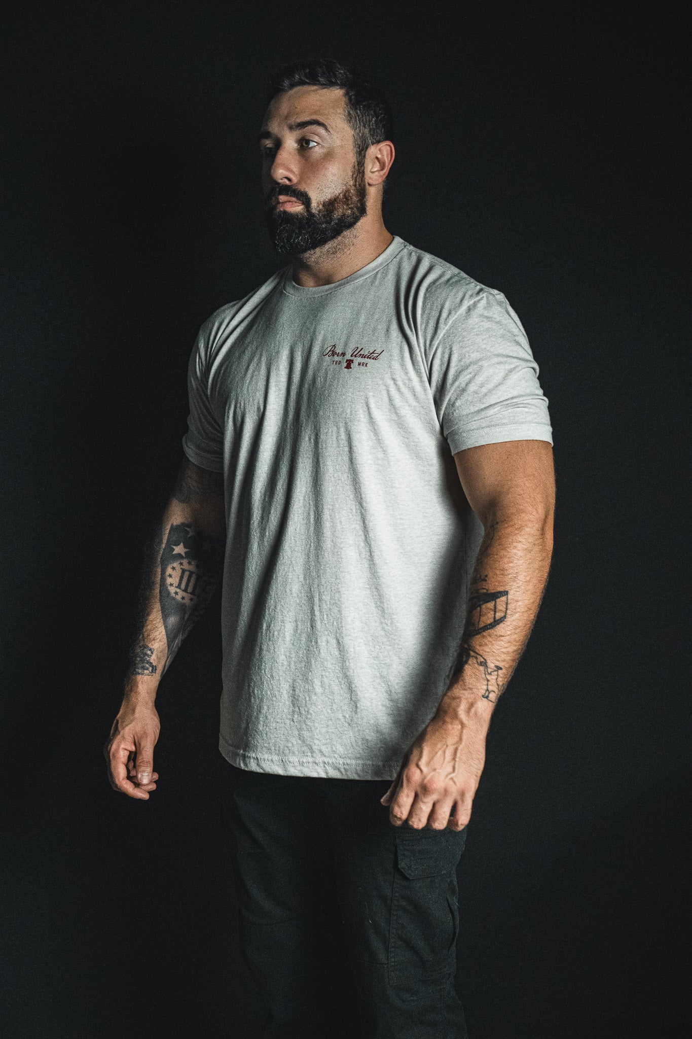 A bearded, tattooed man stands against a dark background, gazing thoughtfully to the side as he wears the “Freedom Not Silenced” light gray t-shirt and black pants from Born United Private Label.