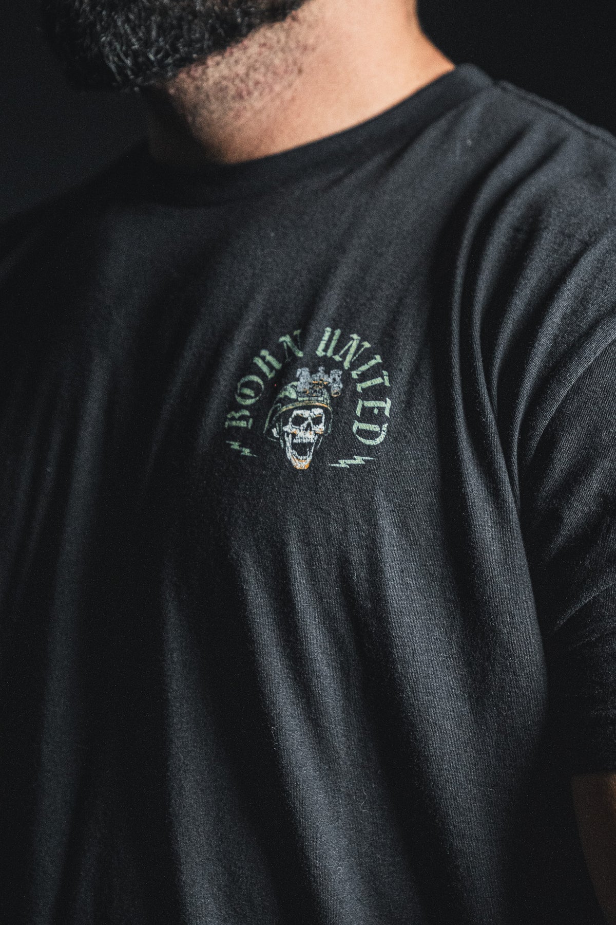 A person wearing a black T-shirt from the Born United Private Label, featuring a small skull graphic and the product name “Compliant We Fall” printed on the chest, along with an inspirational quote: “Defiant we Stand.”.