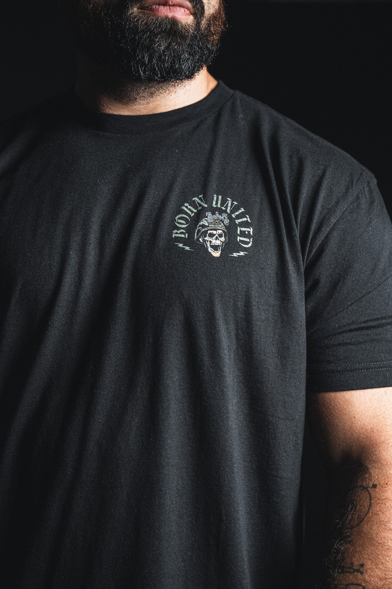 A man with a beard wearing a black t-shirt from Born United Private Label, featuring a small skull graphic and the “Compliant We Fall” text on the upper left chest, paired with the bold phrase “Defiant we Stand” for an added touch of inspiration.