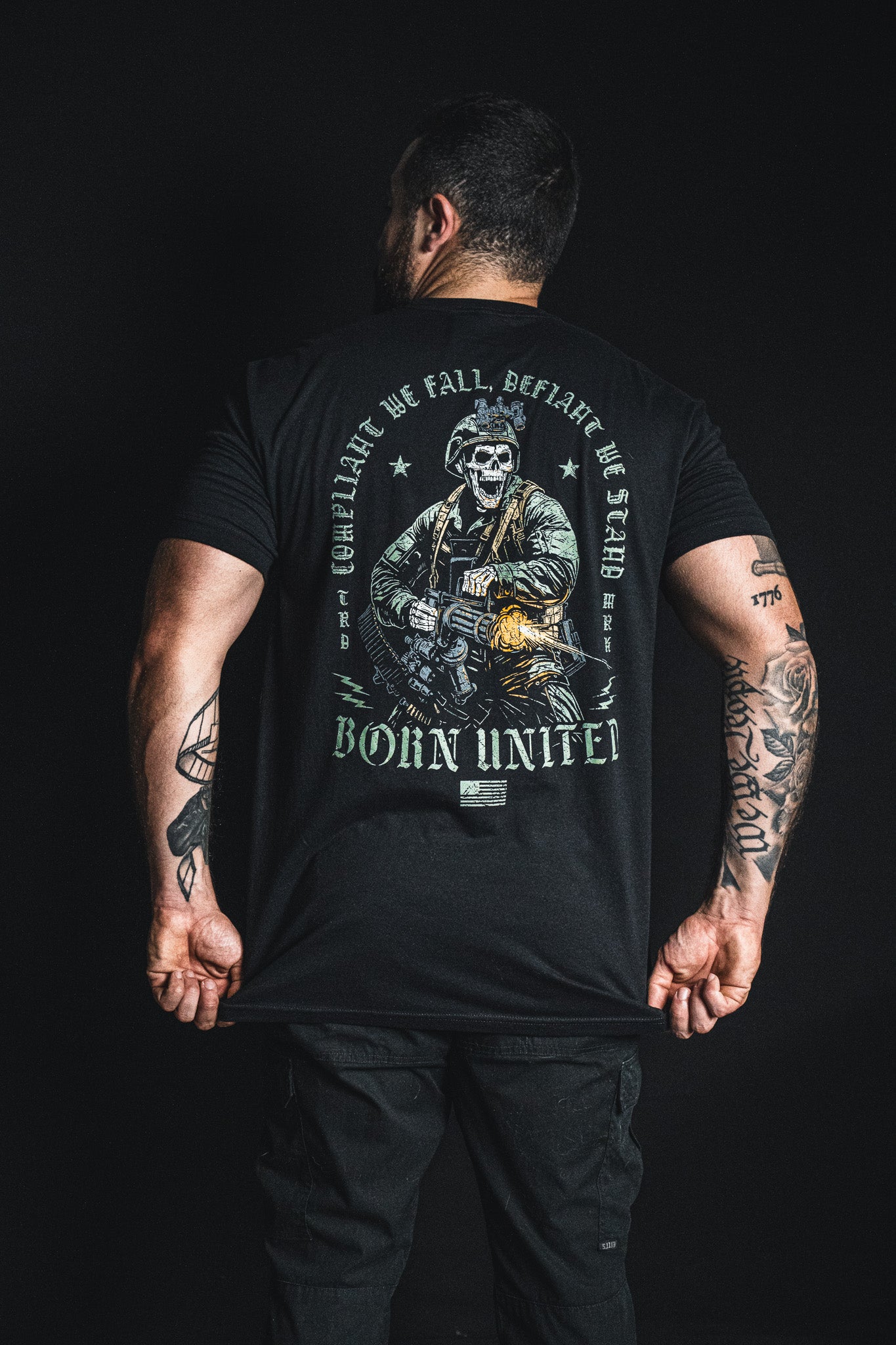 A man faces away, wearing a black Born United Private Label shirt from the “Compliant We Fall” collection. The back features a powerful graphic of a soldier and the bold text: “Born United and Together, Compliant we Fall, Defiant we Stand,” all set against a dark background.