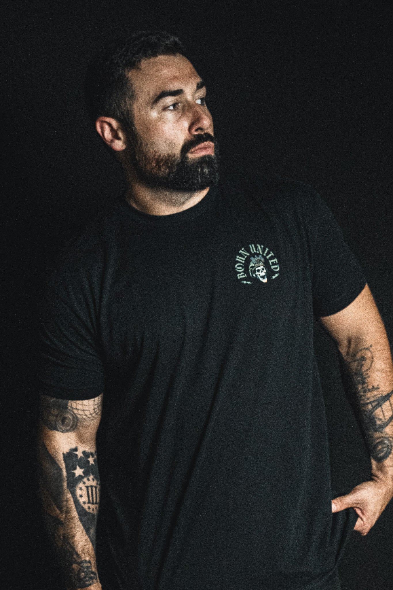 A man with tattoos and a beard stands against a dark background, wearing a black t-shirt from Born United Private Label featuring a small circular emblem on the left chest that reads “Compliant We Fall.”.