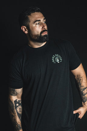 A man with tattoos and a beard stands against a dark background, wearing a black t-shirt from Born United Private Label featuring a small circular emblem on the left chest that reads “Compliant We Fall.”.