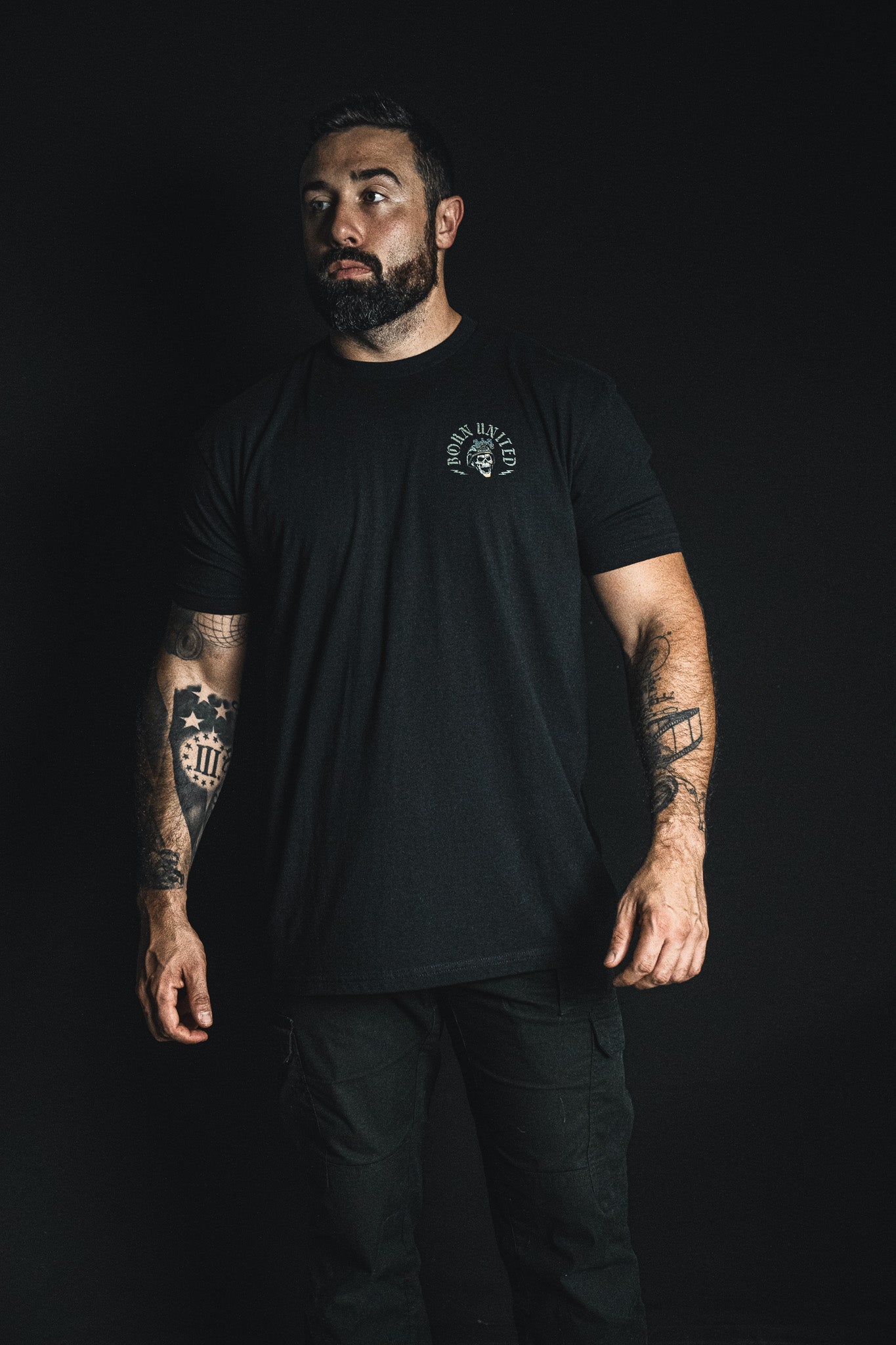 A man with tattoos on both arms stands against a black background, wearing a black t-shirt and pants from Born United Private Label’s “Compliant We Fall” collection, gazing slightly to the side—a striking embodiment of the collection’s bold message.
