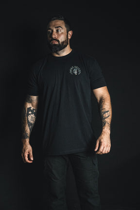 A man with tattoos on both arms stands against a black background, wearing a black t-shirt and pants from Born United Private Label’s “Compliant We Fall” collection, gazing slightly to the side—a striking embodiment of the collection’s bold message.