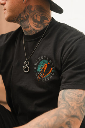 A person with neck and arm tattoos wears the Don't Tread Here Premium Tee by Relentless Betrayal, a black shirt with a green snake design and the brand's name. They also wear a silver necklace featuring a snake-shaped Don't Tread Here pendant.