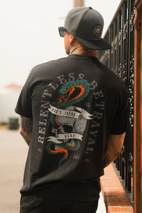 A man in a new backwards cap and the Relentless Betrayal "Don't Tread Here" Premium Tee, featuring a large coiled snake graphic and bold lettering, stands near a black metal fence.
