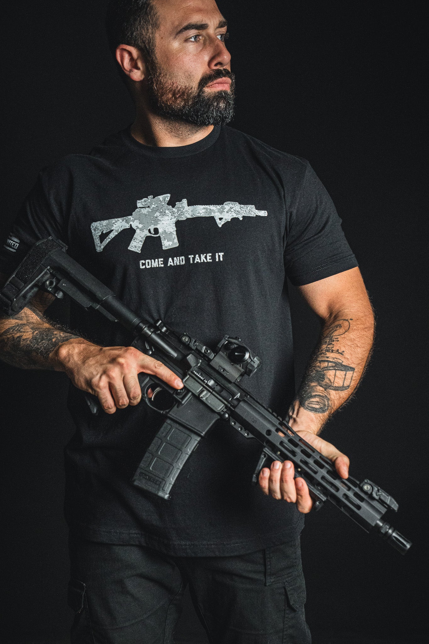 A man holding a rifle wears the Born United Private Label “Come and Take It” patriotic tee, which features a rifle graphic and bold lettering, as he stands against a dark background.