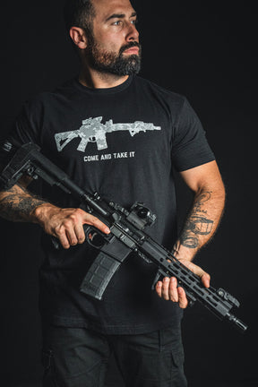 A man holding a rifle wears the Born United Private Label “Come and Take It” patriotic tee, which features a rifle graphic and bold lettering, as he stands against a dark background.