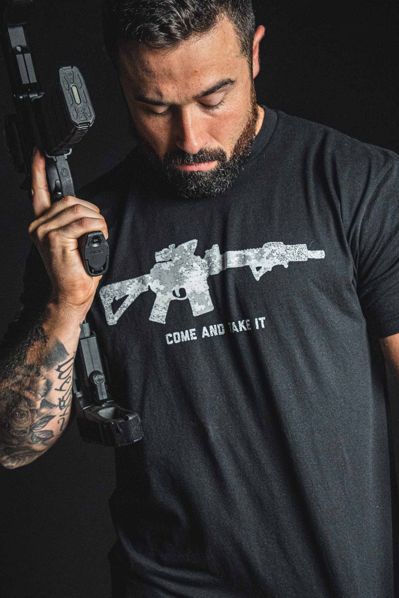 A bearded, tattooed man holds a rifle over his shoulder, wearing the Born United Private Label “Come and Take It” tee—a patriotic shirt featuring an illustrated rifle and bold text that proudly nods to the 2nd Amendment.