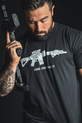 A bearded, tattooed man holds a rifle over his shoulder, wearing the Born United Private Label “Come and Take It” tee—a patriotic shirt featuring an illustrated rifle and bold text that proudly nods to the 2nd Amendment.